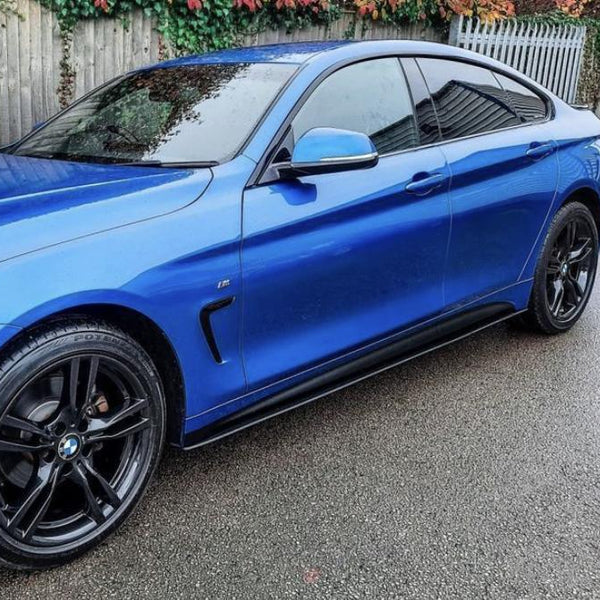 A blue sports car with black wheels, parked on a wet road, is adjacent to a wooden fence and vines with autumn colors cascading over.