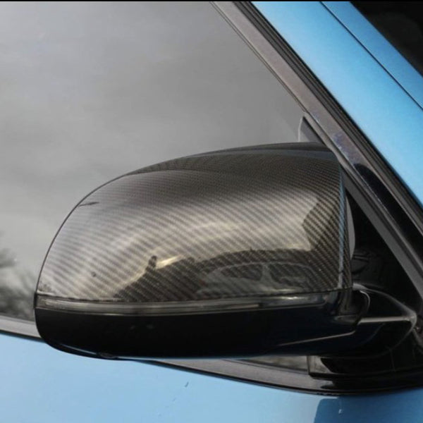 A carbon fiber car side mirror is attached to a shiny blue vehicle. The mirror reflects the cloudy sky and surrounding environment, set against the car's window.