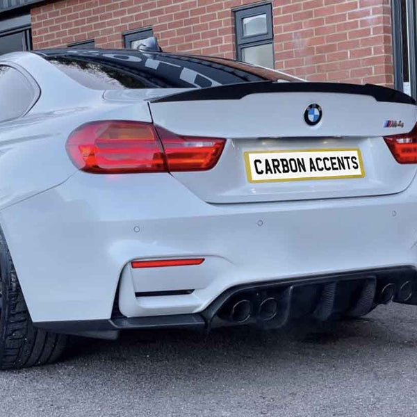 A white BMW car with black rims is parked outside a brick building. The rear license plate reads "CARBON ACCENTS."