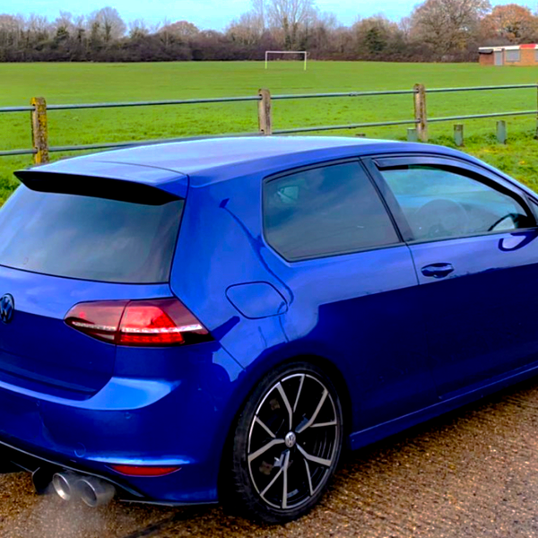 A blue car is parked, emitting exhaust, beside a grassy field with a soccer goal and wooden fencing.