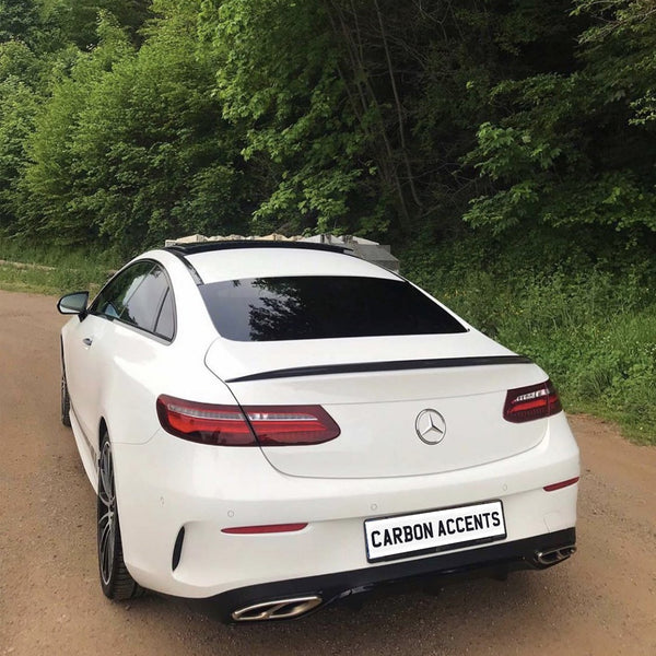 A white Mercedes-Benz coupe is parked on a dirt road, surrounded by lush green trees. The license plate reads "CARBON ACCENTS."
