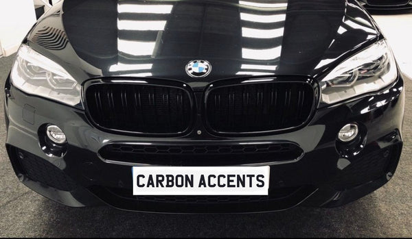 A black BMW car front with a shiny surface reflects bright lights; the grille and headlights are prominent. Text on the license plate reads "CARBON ACCENTS." The car is indoors.