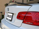 A white BMW features a dark carbon fiber spoiler. The rear view reveals red tail lights and a license plate frame displaying 
