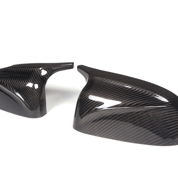 Two carbon fiber side mirror caps, resting on a white background, display a glossy, textured surface with reflective highlights, suggesting a sleek automotive accessory design.
