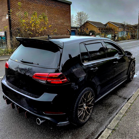 A black hatchback car is parked on a wet street, featuring tinted windows and dual exhaust pipes. It is adjacent to a brick building and some bushes under an overcast sky.