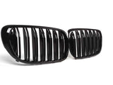 Two glossy black car grilles with vertical slats are positioned side by side, resting on a clean, white surface.