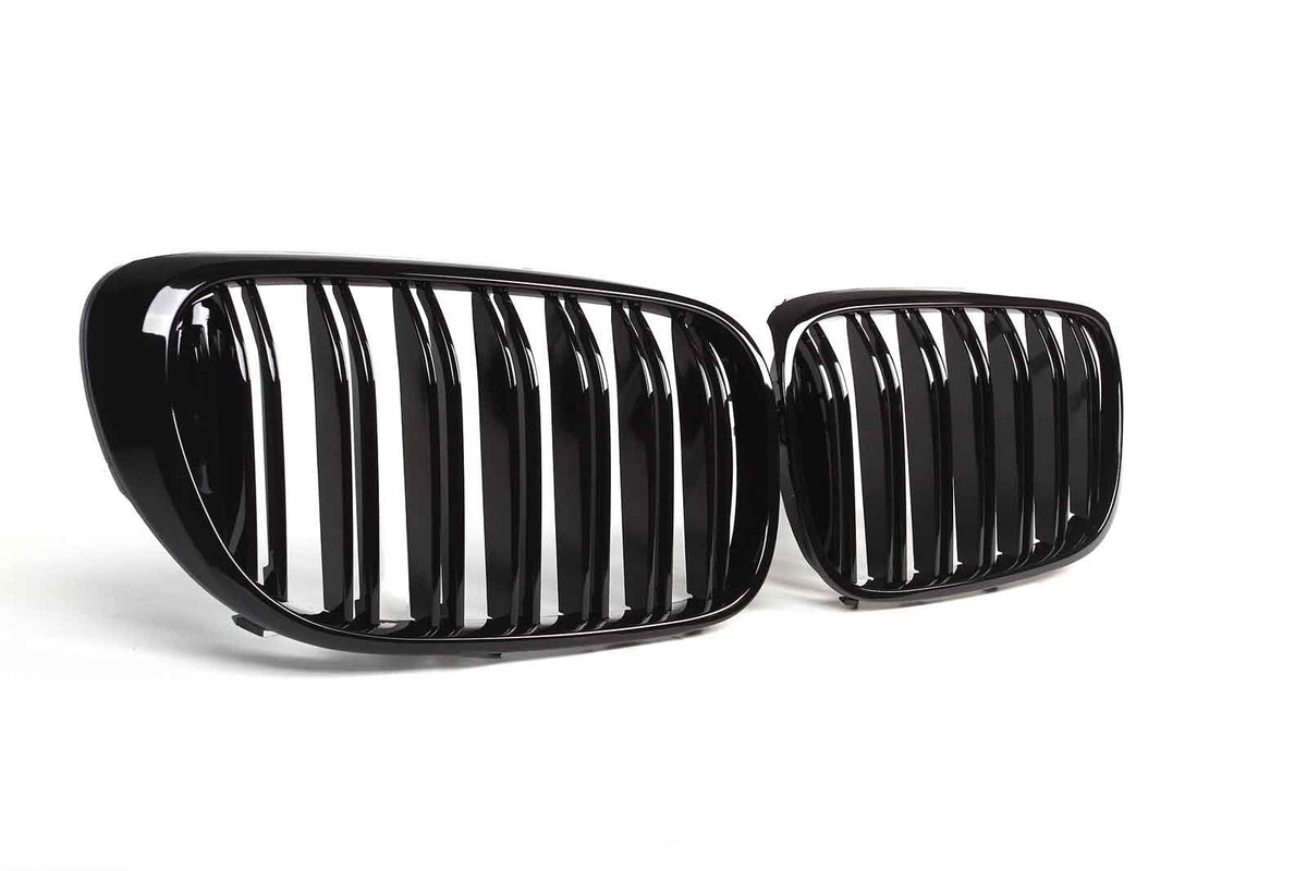 Two glossy black car grilles with vertical slats are positioned side by side, resting on a clean, white surface.