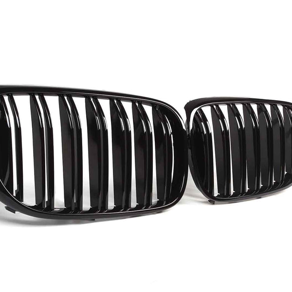 Two glossy black car grilles with vertical slats are positioned side by side, resting on a clean, white surface.