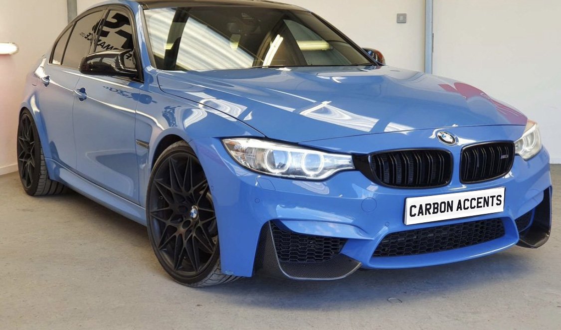 Blue sports car, parked indoors, features custom black wheels and a front license plate reading "CARBON ACCENTS." The setting is a garage with plain walls and reflective lighting.