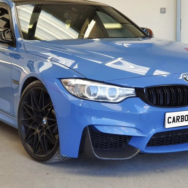 Blue sports car, parked indoors, features custom black wheels and a front license plate reading "CARBON ACCENTS." The setting is a garage with plain walls and reflective lighting.