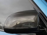 A carbon fiber car side mirror reflects its surroundings, attached to a light blue vehicle, set against a cloudy sky in the background.