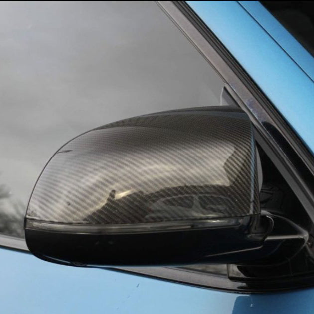 A carbon fiber car side mirror reflects its surroundings, attached to a light blue vehicle, set against a cloudy sky in the background.