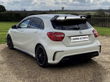 A white hatchback car parked on a gravel driveway, featuring a prominent rear spoiler. The background shows a grassy field and cloudy sky. Text on the license plate reads, 
