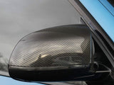 A carbon fiber car side mirror reflects light, attached to a blue vehicle door, against a backdrop of a cloudy sky and faint reflections of surrounding trees.