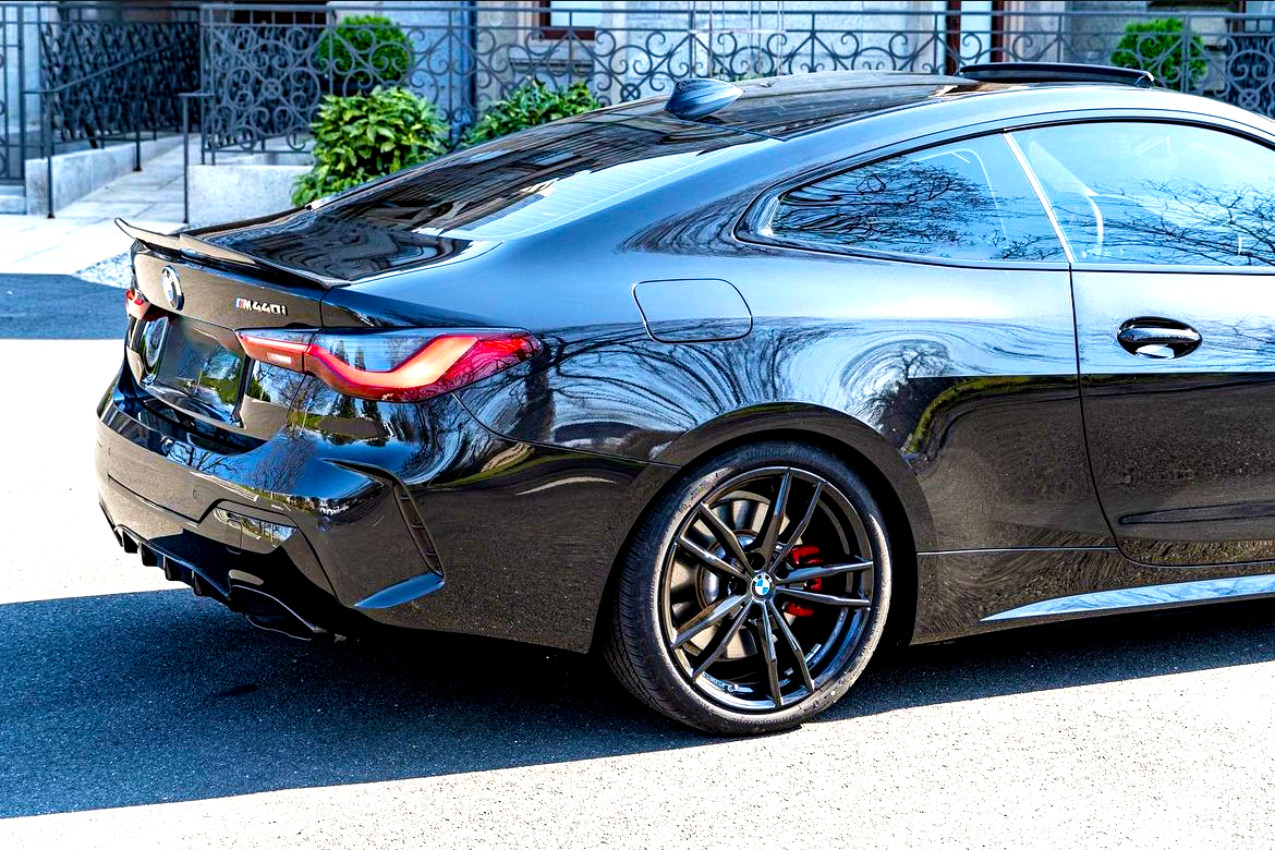 A sleek black BMW car is parked on a sunlit street, showcasing red brake calipers and reflective bodywork. Text reads "M440i." In the background, there are decorative railings and greenery.