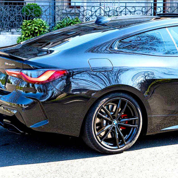 A sleek black BMW car is parked on a sunlit street, showcasing red brake calipers and reflective bodywork. Text reads "M440i." In the background, there are decorative railings and greenery.