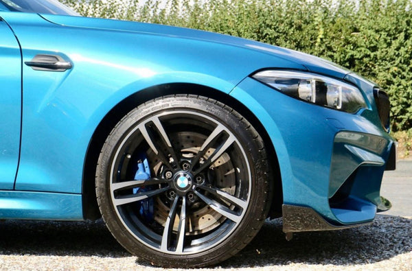 A blue car is parked on gravel, showing its front wheel and headlight. The wheel features a five-spoke design and a visible brake disc. Behind, green shrubs line the background.