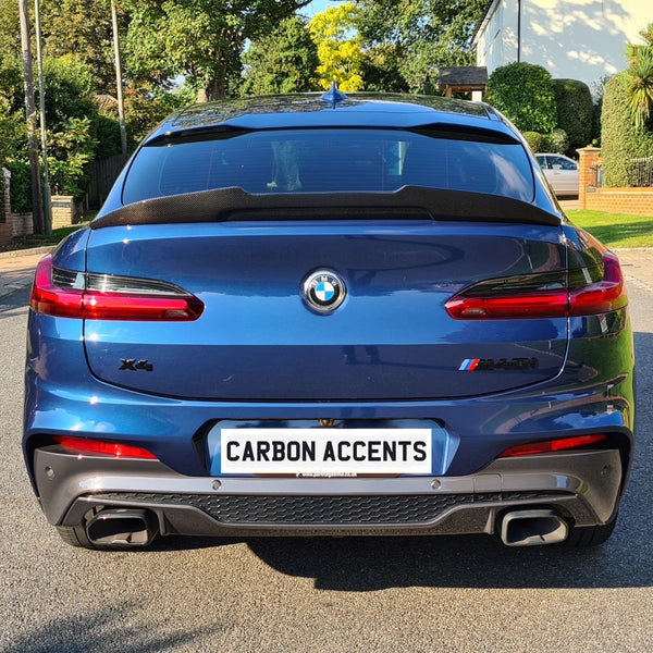 A blue BMW X4 SUV is parked stationary on a sunny suburban street, featuring the license plate text "CARBON ACCENTS." Surrounding are trees and a brick house.