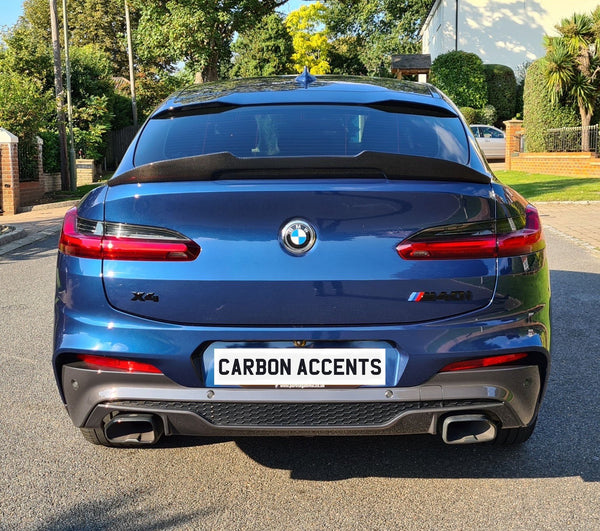 A blue BMW X4 SUV is parked stationary on a sunny suburban street, featuring the license plate text "CARBON ACCENTS." Surrounding are trees and a brick house.