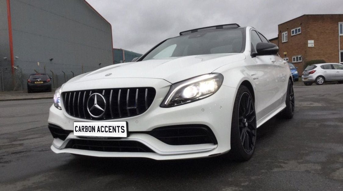 A white car with bright headlights is parked on a wet urban street under an overcast sky. Its license plate reads "CARBON ACCENTS." Industrial buildings surround the scene.