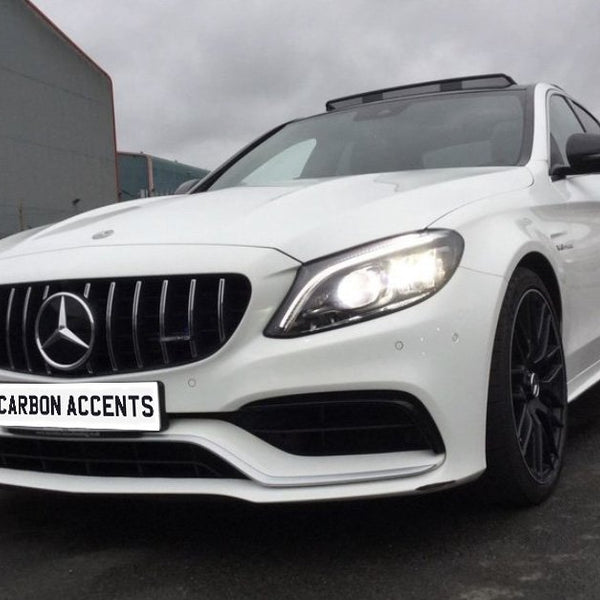 A white car with bright headlights is parked on a wet urban street under an overcast sky. Its license plate reads "CARBON ACCENTS." Industrial buildings surround the scene.