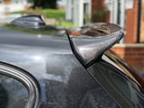 A carbon fiber spoiler is attached to a car's rear in a residential area, reflecting sunlight. A window edge and blurred buildings are visible in the background.