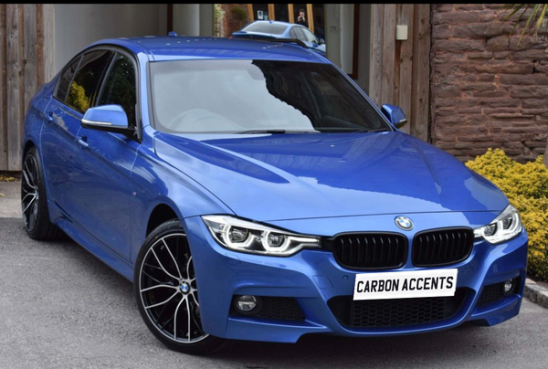 Blue BMW car parked outside a wooden building, featuring black rims and a license plate that reads "CARBON ACCENTS," with bushes and rock wall in the background.