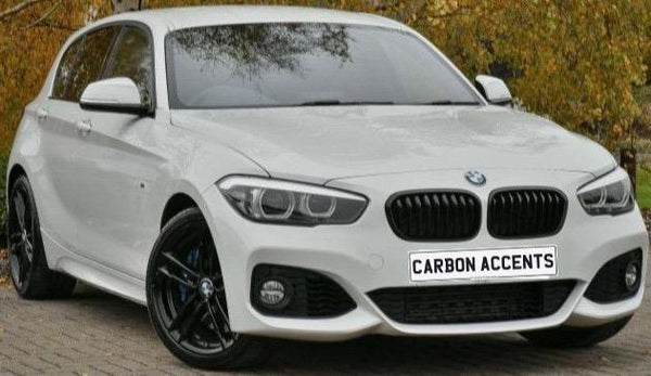 A sleek white sedan with black wheels parked on a brick pavement. Trees with autumn leaves stand in the background. The license plate reads "CARBON ACCENTS."