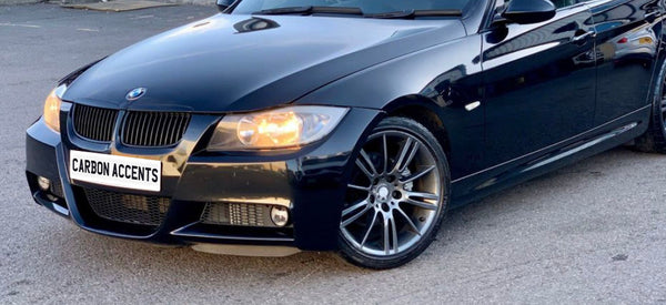 A sleek black car is parked on a street, with its headlights on. The vehicle features alloy wheels and a front license plate that reads "CARBON ACCENTS."