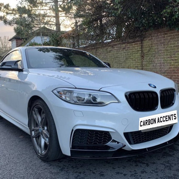 A white BMW parked on a quiet street, with trees and a brick wall in the background. The license plate reads "CARBON ACCENTS."