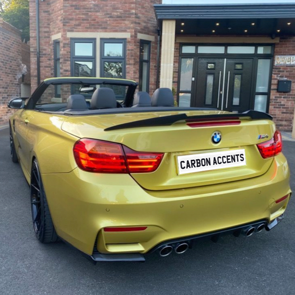 A yellow BMW convertible is parked on a driveway with its top down. The license plate reads "CARBON ACCENTS." A brick building with large windows and a door is in the background.
