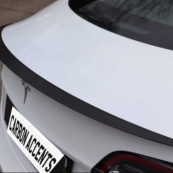 A white car features a sleek, black carbon fiber spoiler on its trunk. The car is parked outdoors, displaying a logo and a license plate reading "CARBON ACCENTS."