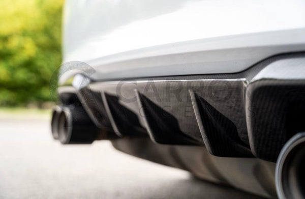 Car rear diffuser with exhaust pipes, surrounded by blurred greenery and pavement. Text reads: "CARBON ACCENTS," faintly visible over the image in gray.