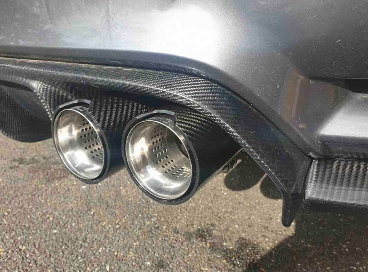 Two shiny metallic exhaust pipes are fixed onto a car's carbon fiber rear diffuser, with a textured road surface visible below in sunlight.