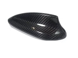 Carbon fiber car side mirror cover, sleek and glossy, resting on a plain white background. The cover exhibits a distinct woven pattern and aerodynamic design, emphasizing high-performance aesthetics.