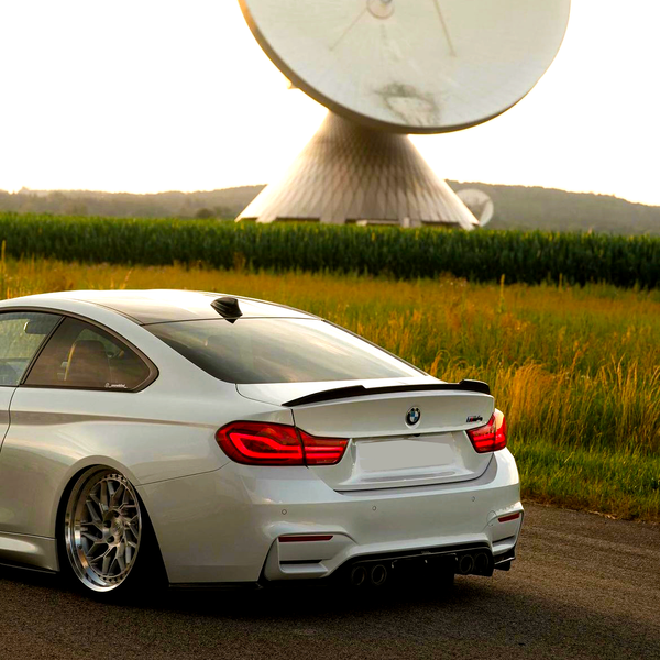 A white car with glossy rims is parked on a road near tall grass, while a large satellite dish looms in the background against a sunset sky.