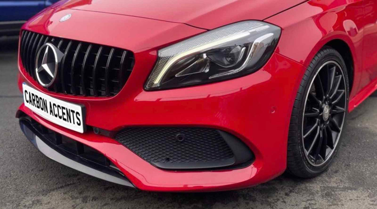 Red car with prominent front grille and sleek headlight, parked on a street. License plate reads "CARBON ACCENTS." A black tire and rim are visible in the foreground.