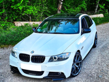 A white BMW car is parked on a gravel path against a backdrop of lush greenery, with sunlight filtering through the trees.