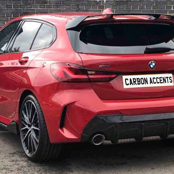 A red BMW hatchback, parked by a brick wall, displays "CARBON ACCENTS" on the license plate. The vehicle is stationary, showcasing sleek, modern design and large wheels.