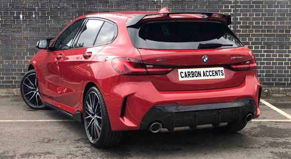 A red BMW hatchback, parked by a brick wall, displays "CARBON ACCENTS" on the license plate. The vehicle is stationary, showcasing sleek, modern design and large wheels.