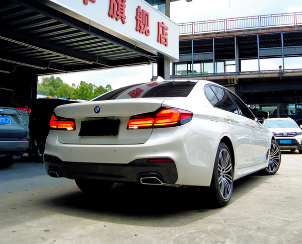 A white BMW with illuminated rear lights is parked under a building with Chinese signage, surrounded by other parked cars on a sunny day.