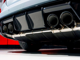 Dual exhaust pipes extend from a car's rear, surrounded by a carbon fiber diffuser. The vehicle is parked on a smooth surface, with a red wall in the background.