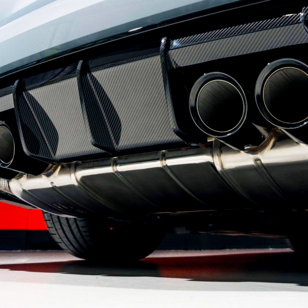 Dual exhaust pipes extend from a car's rear, surrounded by a carbon fiber diffuser. The vehicle is parked on a smooth surface, with a red wall in the background.