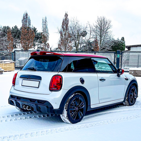 A white car with a red roof is parked on snow, leaving tire tracks. It's in a fenced area with trees and a building in the background.