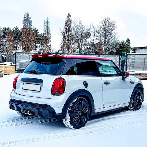 A white car with a red roof is parked on snow, leaving tire tracks. It's in a fenced area with trees and a building in the background.