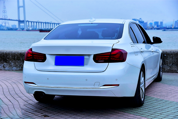 A white car is parked on a tiled waterfront promenade, with a bridge and city skyline visible across the water. The car's license plate is blue.