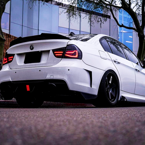 White sports car parked, facing left, with illuminated tail lights. It is on a street with brick and glass buildings, and trees are nearby, creating an urban setting.