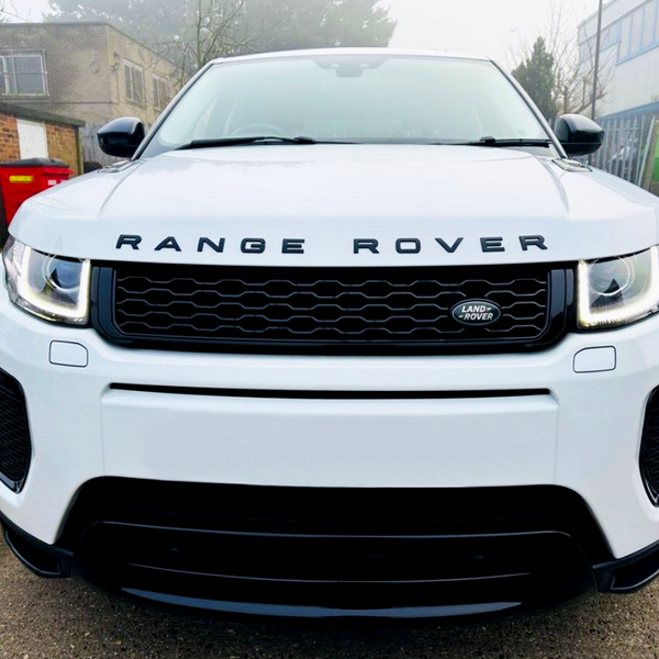 A white Range Rover, parked on a driveway, features a prominent front grille with the "Land Rover" logo. Overcast sky and industrial buildings set the backdrop.