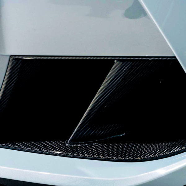 A car's carbon fiber air intake vent features a slatted design, positioned on the vehicle's front bumper. The glossy finish contrasts with the surrounding white bodywork, suggesting a sporty aesthetic.