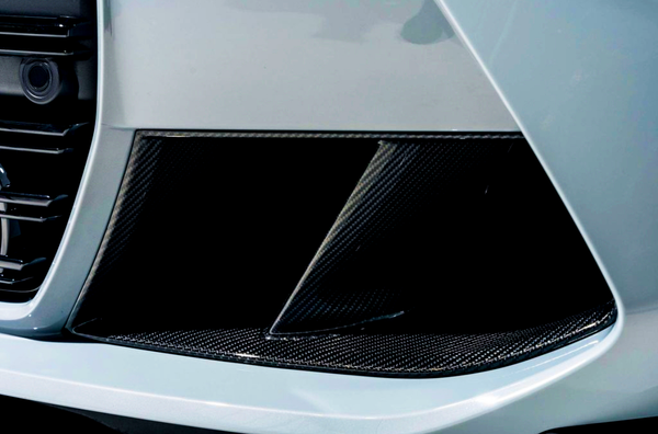 A car's carbon fiber air intake vent features a slatted design, positioned on the vehicle's front bumper. The glossy finish contrasts with the surrounding white bodywork, suggesting a sporty aesthetic.